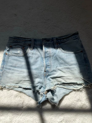 Ljusblåa högmidjade jeansshorts - Säljer ett par ljusblåa högmidjade jeansshorts med fransiga kanter och klassisk femficksdesign. Shortsen har knappar i olika färger för en unik touch. 