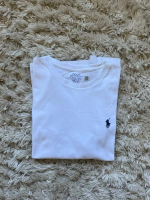 Vit slim fit t-shirt från Polo Ralph Lauren - Klassisk vit  custom slim fit t-shirt från Polo Ralph Lauren med rund halsringning och den ikoniska mörkblå logotypen broderad på bröstet. Tillverkad i mjuk bomull för en skön känsla och perfekt passform. Enkel och stilren design som funkar till allt.