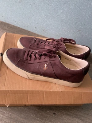 Vinröda Polo Ralph Lauren sneakers - Snygga vinröda sneakers från Polo Ralph Lauren med klassisk låg profil och vit sula. Skorna har canvasmaterial, matchande vinröda skosnören och den ikoniska broderade loggan på sidan. Perfekta för en avslappnad och trendig stil.