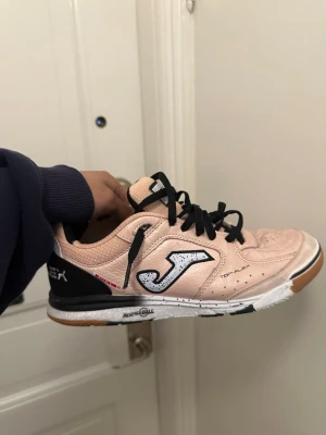 Joma Top-Flex rosa rebound - Säljer ett par Joma Top-Flex i storlek 40.5 i ljusrosa med svart och vitt Joma-märke på sidan. Skorna har en gummisula för inomhusspel och är tillverkade i syntetmaterial och mockaimitation. Ena skon har svarta snören och den andra har ljusrosa snören för en unik look.