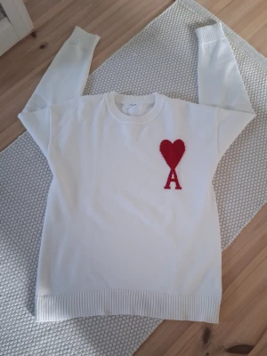 Ami Paris tröja - Vit stickad tröja från Ami Paris med röd hjärtdetalj och bokstaven A på bröstet. Tröjan har rund halsringning, ribbade muddar och nederkant. Perfekt för dig som gillar stilrena plagg med en twist. Storlek M men passar även S. Inga defekter alls. Tags är avklippta, men medföljer. Hör av dig vid minsta fundering eller ifall du vill ha fler bilder! 