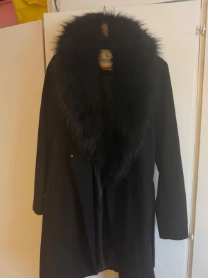 Svart kappa med fuskpäls från Chiara Forthi - Stilren svart kappa från Chiara Forthi med stor, fluffig fuskpäls runt kragen. Kappan har rak passform och är helsvart, vilket ger en lyxig och trendig look. Perfekt för dig som vill sticka ut med en elegant vibe.