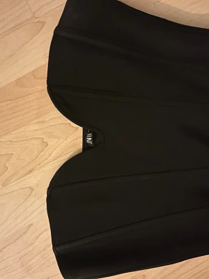 Svart korsettopp från Zara - Säljer en svart korsettopp från Zara med en snygg hjärtformad överkant. Toppen är axelbandslös och har markerade sömmar för en strukturerad look. Perfekt att styla med höga jeans eller kjol för en trendig outfit.