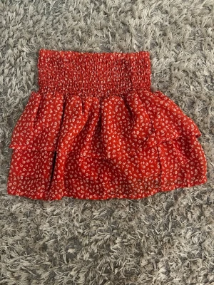 Röd blommig volangkjol - Supersöt röd kortkjol med små vita blommor och volang. Kjolen har en smockad midja som gör den bekväm och sitter snyggt. Perfekt för varma dagar och ger en riktigt somrig vibe. Lappen är lös