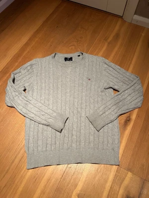 Ljusgrå kabelstickad tröja från Gant - Säljer en ljusgrå kabelstickad tröja från Gant i premium bomull. Tröjan har rund hals, ribbade muddar och diskret Gant-logo på bröstet. Perfekt för lager-på-lager och passar till både jeans och chinos. 
