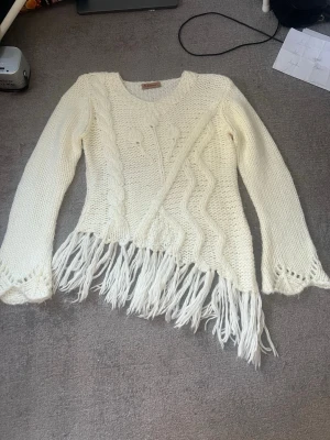 Vit stickad tröja med fransar Zephirus - Säljer en vit stickad tröja från Zephirus med långa ärmar, dekorativa flätmönster och vågiga detaljer framtill. Tröjan har en unik design med fransar längs nederkanten och är gjord i ett tjockare garn som känns mjukt och mysigt.