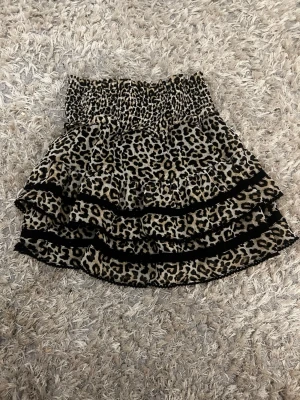 Leopardmönstrad volangkjol - Kort kjol med leopardmönster och volanger. Kjolen har bred resår i midjan och svarta spetsband längs volangerna. Perfekt för dig som vill sticka ut med en trendig och djurinspirerad look.