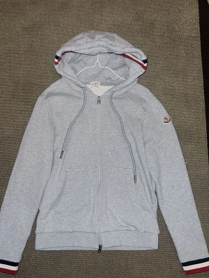 Gråå Moncler kofta - Snygg ljusgrå hoodie från Moncler med dragkedja, huva med dragsko och fickor framtill. Detaljer i rött, vitt och blått på huva och ärmslut samt Moncler-logga på ärmen. Tillverkad i mjukt bomullsmaterial för en skön känsla. ‼️NFC och QR kod funkar‼️
