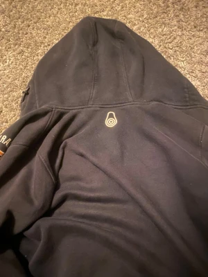 Svart Sail Racing hoodie med dragkedja - Svart hoodie från Sail Racing med dragkedja och huva. Insidan av huvan är orange, och på bröstet finns texten 'SAILRACING 77' i vitt och gult. Mjuk bomullskänsla och diskret Sail Racing-logga på ryggen. Perfekt för en sportig och avslappnad stil.