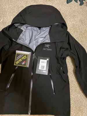 Svart Arc'teryx Beta LT vindjacka - Arc'teryx Beta LT vindjacka i svart med GORE-TEX-material. Jackan har justerbar huva, vattentäta dragkedjor och två sidofickor. Perfekt för blåsiga och regniga dagar. Minimalistisk design med Arc'teryx-logga på bröstet.