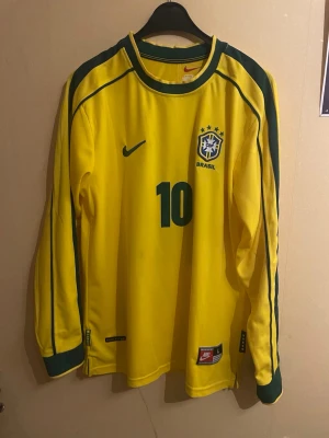 Rivaldo - Brasilien #10 - Säljer en klassisk Brasilien landslagströja med Rivaldo #10, långärmad i originaldesign från Nike. Ikonisk gul tröja med gröna detaljer, CBF-emblemet med stjärnor och det tidstypiska snittet som användes under Rivaldos storhetstid. Ett riktigt samlarobjekt och en dröm för alla Brasilien-fans och fotbollsentusiaster. Tröjan är i fint skick med tryck och detaljer väl bevarade. Perfekt både för samling, nostalgibruk eller som statement-plagg.