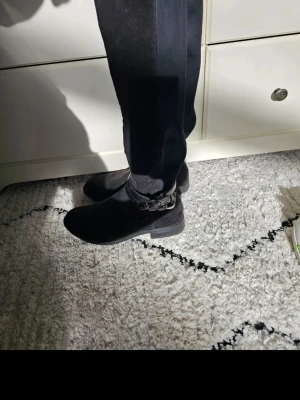 Svarta knähöga stövlar med kedjedetalj - Säljer ett par svarta knähöga stövlar med snygg kedjedetalj runt ankeln och dragkedja på insidan. Stövlarna har en smal passform och låg klack. Materialet ser ut att vara mocka eller mockaimitation med stretchpanel längs skaftet för extra komfort.
