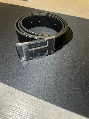 Svart bälte skärp med H-spänne - Stilrent svart skärp med H-formad metallspänne. Skärpet har vita kontrastsömmar längs kanterna och är tillverkat i skinn. Perfekt accessoar för att lyfta din outfit med en lyxig touch.