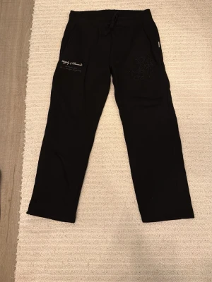 Supply and demand byxor  - Säljer ett par svarta mjukisbyxor med broderad detalj och tryck på vänster sida. Byxorna har raka ben, dragsko i midjan och sidofickor. Perfekta för chill dagar eller när du vill vara bekväm. Materialet känns mjukt och skönt mot huden.