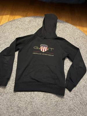 Svart hoodie från Gant med logga - Svart hoodie från Gant med stor huva och magficka. På bröstet finns Gant-logga och texten 'American Sportswear' i guld och vitt, samt ett rött och vitt sköldmärke. Tillverkad i mjukt bomullsmaterial, perfekt för en avslappnad och sportig stil.