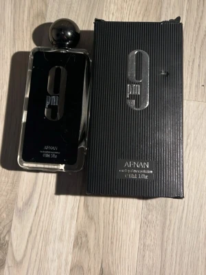 Afnan 9PM Eau de Parfum  - Afnan 9PM Eau de Parfum är en stilren herrdoft i en rektangulär glasflaska med svart etikett och silverdetaljer. Flaskan har en rund, svart kork och kommer i en matchande svart kartong med silvertext. Volymen o ml