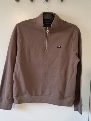 Brun halvzip sweatshirt från Lexington storlek 170 - Snygg brun sweatshirt från Lexington med halv dragkedja och liten logotyp på bröstet. Tröjan har hög krage och ribbade muddar vid ärmslut och nederkant. Perfekt för en avslappnad och stilren look.