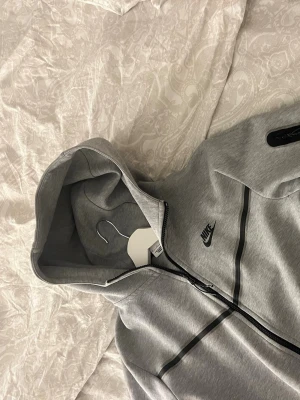 Grå Nike hoodie med dragkedja - Snygg grå hoodie från Nike med svart logga på bröstet och svarta detaljer längs dragkedjan. Hoodien har huva och hel dragkedja framtill, samt är tillverkad i mjukt material som passar perfekt till en avslappnad stil.