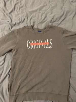 Grön sweatshirt med tryck - Säljer en grön sweatshirt med texten 'ORIGINALS GENERATION' i vitt och rött framtill. Tröjan har rund halsringning, långa ärmar och ribbade muddar vid ärmslut och nederkant. Perfekt för en avslappnad och trendig stil.
