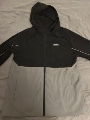 Svart och grå Mckenzie vindjacka - Snygg tvåfärgad vindjacka från Mckenzie i svart och ljusgrått. Jackan har huva med dragsko, hel dragkedja framtill, två sidofickor och en bröstficka med dragkedja. Detaljer på ärmarna och stilren design. Perfekt för blåsiga dagar.