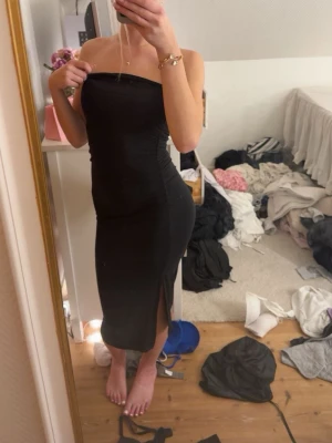 Svart axelbandslös klänning med slits - Säljer en svart, axelbandslös klänning i figurnära modell. Klänningen har en snygg slits framtill och är i en längre längd som ger en elegant siluett. Perfekt för dig som vill ha en stilren och enkel look.