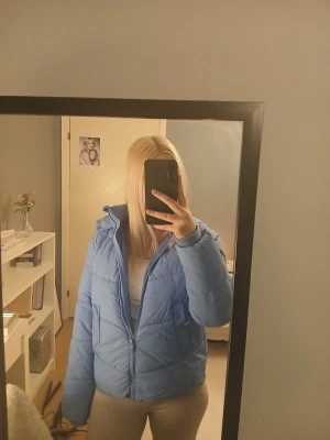 Ljusblå pufferjacka med huva - Säljer en ljusblå pufferjacka med dragkedja och huva. Jackan har en kortare modell och quiltad design som ger en snygg och modern look. Perfekt för kalla vinterdagar när du vill hålla dig varm och stilren. Andvänt knappt. 