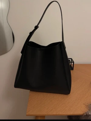 Svart väska från Vero Moda  - Stilren svart väska med justerbart axelband och minimalistisk design. Väskan har en rymlig insida utan synliga loggor eller detaljer. Perfekt för dig som gillar enkel och modern stil.