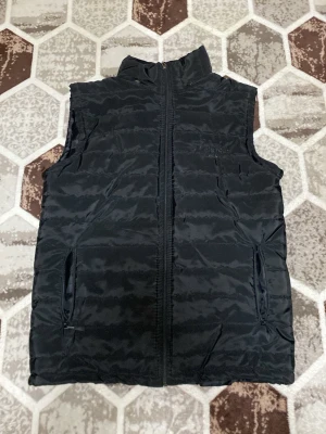 Svart dunväst från Columbia Titanium - Stilren svart dunväst från Columbia Titanium med hög krage och dragkedja framtill. Västen har två praktiska fickor med dragkedja och diskret logga på bröstet. Perfekt för lager-på-lager och outdoor-looks.