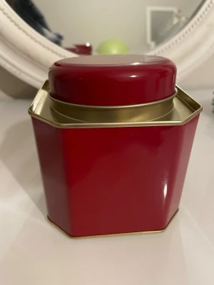 Röd plåtburk med lock - Snygg röd plåtburk med guldiga detaljer och lock. Burken är sexkantig och passar perfekt för att förvara te, kaffe eller småprylar. Stilren design som gör sig bra i köket eller på skrivbordet.
