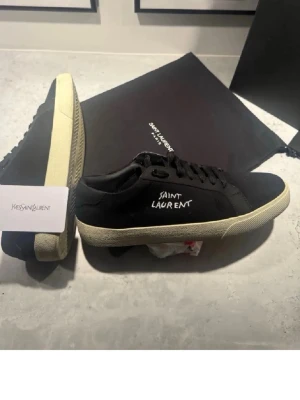 Svarta Saint Laurent sneakers - Snygga svarta sneakers från Saint Laurent med vit textlogga på sidan och kontrasterande vit sula. Klassisk låg modell med svarta skosnören och stilren design. Perfekta för dig som gillar exklusiva streetwear-vibbar.
