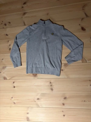 Grå halvzip tröja från Lyle & Scott - Snygg grå tröja från Lyle & Scott med halv dragkedja och klassisk gul logga på bröstet. Tröjan har ribbade muddar och hög krage, perfekt för lager på lager. Tillverkad i merino ull mix.