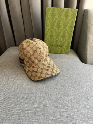 Beige keps från Gucci med GG-mönster - Snygg beige keps från Gucci med klassiskt GG-monogram över hela kepsen. Kepsen har böjd skärm och en ikonisk grön-röd rand på sidan. Tillverkad i canvasmaterial med bruna detaljer. Perfekt accessoar för en trendig look.