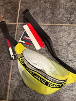 Gul shoppingväska från Tommy Jeans - Cool shoppingväska från Tommy Jeans i en fräsch gul färg med svart justerbart band. Väskan har en dragkedja framtill och ett tydligt Tommy Jeans-tryck längs dragkedjan. Perfekt för att bära dina grejer med stil.