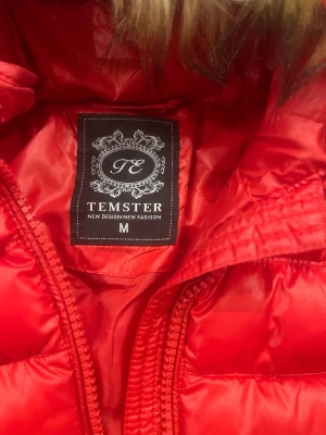 Röd puffjacka med fuskpäls från Temster - Snygg röd puffjacka från Temster med huva och fuskpäls. Jackan har två vertikala dragkedjefickor framtill och quiltad design. Perfekt för kalla dagar och ger en trendig look.