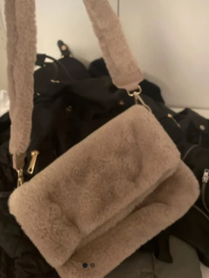 Beige fluffig axelväska - Supersöt beige axelväska i mjukt fuskpälsmaterial med bred fluffig axelrem. Väskan har en rektangulär form och stängs med lock. Perfekt för dig som vill ha en mysig och trendig accessoar till din outfit.