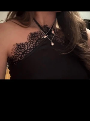 Svart spetsdetaljerad linnetopp - Snygg svart linnetopp med tunna axelband och dekorativa spetsdetaljer längs urringningen. Toppen har en halterneck-modell och är tillverkad i ett mjukt, glansigt material som ger en elegant look. Perfekt att styla med smycken för en trendig outfit. Högt pris pga intressekoll