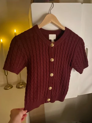 Vinröd kabelstickad kofta kort ärm - Kortärmad vinröd kofta med kabelstickat mönster och guldfärgade knappar framtill. Rund halsringning och ribbade muddar vid ärmslut och nederkant. Perfekt för lager på lager och en snygg detalj till din outfit.