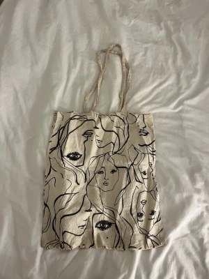 Beige tygkasse med konstnärligt ansikte - Trendig shoppingväska i beige tyg med svart konstnärligt ansiktsmönster. Väskan är välanvänd
