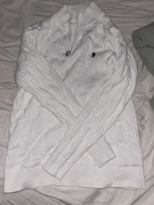 Vit kabelstickad tröja Polo Ralph Lauren - Kabelstickad vit tröja från Polo Ralph Lauren med halv dragkedja och broderad logga på bröstet. Tröjan har hög krage, långa ärmar och ribbade muddar. Perfekt för en clean och klassisk look. Storlek S passar xs
