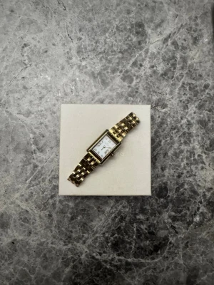 Guldfärgad rektangulär klocka - Stilren klocka med rektangulär vit urtavla och guldfärgad länk i metall. Klassisk design med tydliga svarta siffror och visare. Perfekt accessoar för dig som gillar tidlös och elegant stil.