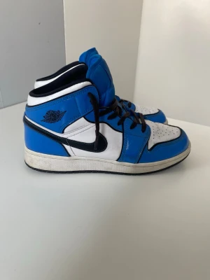 Nike Air Jordan 1 High - Nike Air Jordan 1 High i blå, vit och svart färgkombination. Klassisk high-top-modell med Air Jordan-logga och svart Swoosh. Skorna är i fint begagnat skick med normalt slitage, främst på sulorna samt mindre märken på lädret. Inga större skador.