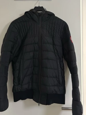 Svart dunjacka med huva Canada Goose - Snygg svart dunjacka från Canada Goose med huva och dragkedja framtill. Perfekt för kalla vinterdagar och håller dig varm med sin vadderade insida. Lite sliten i armarna (kolla bilderna) storlek m/m liten i storlek så passar bra på dom som har xs/s