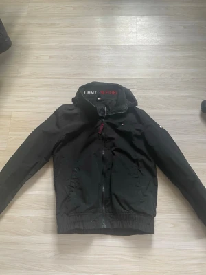 Svart vindjacka från Tommy Hilfiger - Snygg svart vindjacka från Tommy Hilfiger med dragkedja framtill, hög krage med logga och elastiska muddar. Jackan har två sidofickor och är tillverkad i ett lätt, vindtätt material. Perfekt för blåsiga dagar och enkel att matcha med olika outfits.