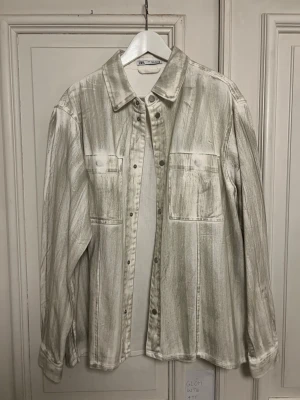 Beige overshirt från Zara - Säljer en beige overshirt från Zara med borstade, nästan penseldrag-liknande detaljer. Skjortan har två stora bröstfickor, knappar framtill och klassisk krage. Materialet känns som bomull eller denim och modellen är rak och avslappnad.