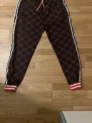 Gucci mjukisbyxor med logomönster - Svarta mjukisbyxor från Gucci med rött GG-logomönster över hela byxan, vita och röda ränder längs sidorna samt mudd vid benslut med röd och vit rand. Resår i midjan med rosa dragsko. Perfekta för en trendig och avslappnad stil.