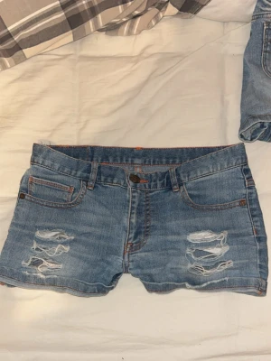 Blå jeansshorts med slitningar - Snygga blå jeansshorts med slitna detaljer framtill och klassisk femficksdesign. Shortsen har låg midja och är tillverkade i bomull med en knapp och dragkedja framtill. Perfekta för sommardagar och en avslappnad stil.