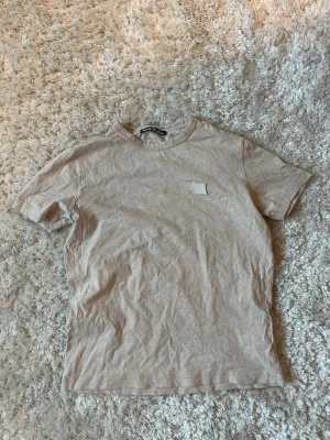 Beige t-shirt från Acne Studios - Säljer en beige t-shirt från Acne Studios med det klassiska broderade Face-logotypen på bröstet. T-shirten har rund hals och korta ärmar, tillverkad i mjuk bomull för en skön känsla. Perfekt basic-plagg till garderoben.