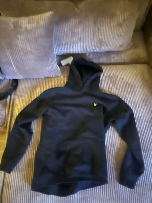 Svart hoodie från Lyle & Scott - Snygg svart hoodie från Lyle & Scott med klassisk gul logga på bröstet. Hoodien har huva och en stor magficka framtill. Tillverkad i mjukt material som känns skönt mot huden. Perfekt för dig som gillar enkel och stilren streetwear.