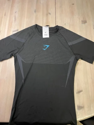 Gymshark onyx svart Compression shirt med lappen kvar - Snygg svart t-shirt från Gymshark med gråa detaljer och ett blått logotryck på bröstet. Tröjan har korta ärmar och är tillverkad i ett stretchigt syntetmaterial som passar perfekt för träning. Modern design med mönstrade partier på både fram- och baksida. / svår att få tag på. Lappen sitter fortfarande på!