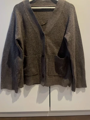 Brun kofta från 157 med fickor - Mysig brun kofta från 157 med v-ringning, stora framfickor och knäppning med tre knappar. Koftan har ribbade muddar vid ärmslut och nederkant, och är gjord i ett mjukt stickat material. Perfekt för lager på lager och chill dagar.